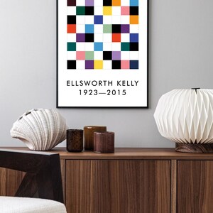 May include: A black framed print with a colorful geometric pattern and the text "ELLSWORTH KELLY 1923-2015". The print is hanging on a light gray wall above a wooden cabinet.