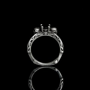 Könnte beinhalten: Ein silberner Ring mit einem schwarzen Edelstein und Totenkopfdetails. Der Ring ist vor einem schwarzen Hintergrund platziert und spiegelt sich in der Oberfläche.