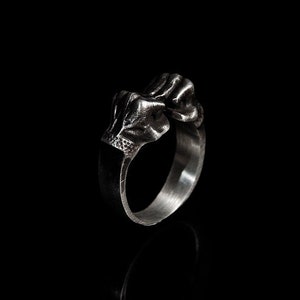 May include: A silver ring with a detailed skull design. The skull is depicted with a clenched fist.