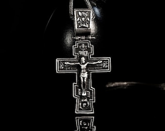 May include: A silver Orthodox cross pendant with intricate details and a depiction of Jesus Christ on the crucifix. The cross is hanging from a black cord.