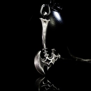 May include: A silver axe pendant with intricate details, hanging from a chain against a black background.