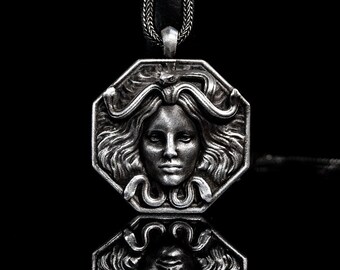 May include: A silver pendant necklace featuring a detailed, octagonal design with a woman's face surrounded by snakes. The pendant is reflected in a black surface.