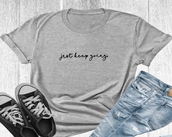 May include: A gray t-shirt with the text "just keep going" in black script lettering. The shirt is laid flat on a wooden surface with a pair of black canvas shoes and blue jeans.