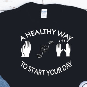 Op de afbeelding: Een zwart T-shirt met witte tekst die "A healthy way to start your day" luidt, met twee witte handen in gebedshouding aan weerszijden van de tekst en een witte afbeelding van een persoon die aan het sporten is in het midden.