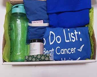 Op de afbeelding: Een blauwe cadeaudoos gevuld met spullen voor iemand die een kankerbehandeling ondergaat. De doos bevat een blauw chemo-poortkussen, een blauwe nekwarmer, een groene waterfles, een klein potje bodycrème, een groen-wit gestreept stofje en een blauw t-shirt met de tekst "Do List: Beat Cancer".
