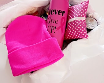 Op de afbeelding: Een roze cadeaudoos gevuld met roze items, waaronder een roze beanie, een roze waterfles met de tekst "Never Give Up!" en een roze lint, en een roze tas met witte stippen.
