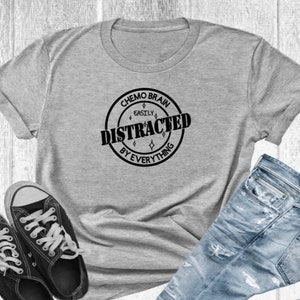 May include: A gray t-shirt with the text "Chemo Brain Easily Distracted By Everything" in a black circular design. The shirt is paired with black sneakers and blue jeans.