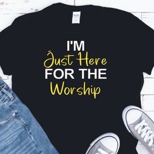 May include: Black t-shirt with white text that reads "I'm Just Here For The Worship" in yellow.