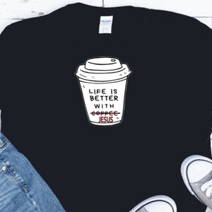 Op de afbeelding: Zwart T-shirt met een witte afbeelding van een koffiebeker. De tekst op de beker luidt "Life is better with Jesus", met "coffee" doorgestreept. Het shirt wordt gecombineerd met een blauwe jeans en grijze sneakers.