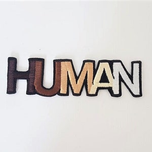 Op de afbeelding: Een zwarte geborduurde patch met het woord "HUMAN" in witte, bruine en beige letters. De letters zijn gelaagd om verschillende huidtinten weer te geven.