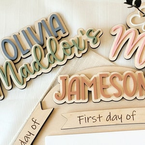 May include: Wooden name plaques with the names Olivia, Maddox, Jameson, Mila and Evelyn. The plaques are in various shades of green, brown, pink and white. The plaques are arranged on a wooden surface with a sign that says "First day of".