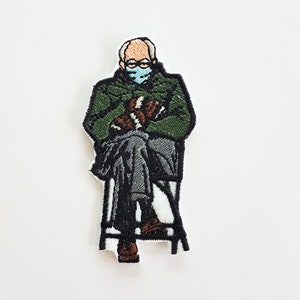 Può includere: Toppa ricamata di una persona che indossa una giacca verde e una maschera facciale blu, seduta su una sedia con le mani in grembo.