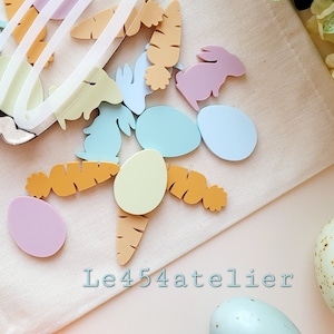 May include: A collection of pastel colored wooden Easter decorations, including eggs, carrots, and bunnies, on a white fabric surface. The text "Le454atelier" is visible in the lower right corner of the image.