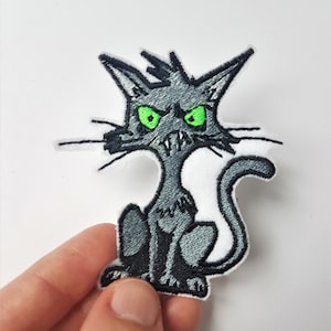 Peut inclure: Un patch brodé gris à repasser représentant un chat de dessin animé avec des yeux verts et une expression boudeuse.