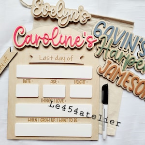 Könnte beinhalten: Holzschild mit weißem Hintergrund und leeren Feldern zum Ausfüllen mit Datum, Alter, Größe, Dingen, die du liebst, und was du werden möchtest, wenn du groß bist. Das Schild hat ein rosa-weißes Namensschild "Caroline's", ein weiß-braunes Namensschild "Evelyn's" und ein blau-grün-braunes Namensschild "Gavin's Harper's Jameson". Das Schild wird mit einem schwarzen Stift und einer Stofftasche geliefert.