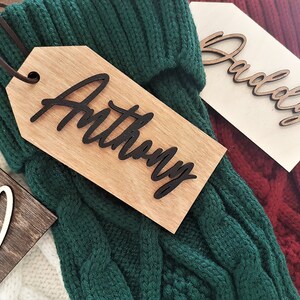 May include: Three wooden gift tags with names written in black script. The tags are attached to green, white, and red knitted stockings.