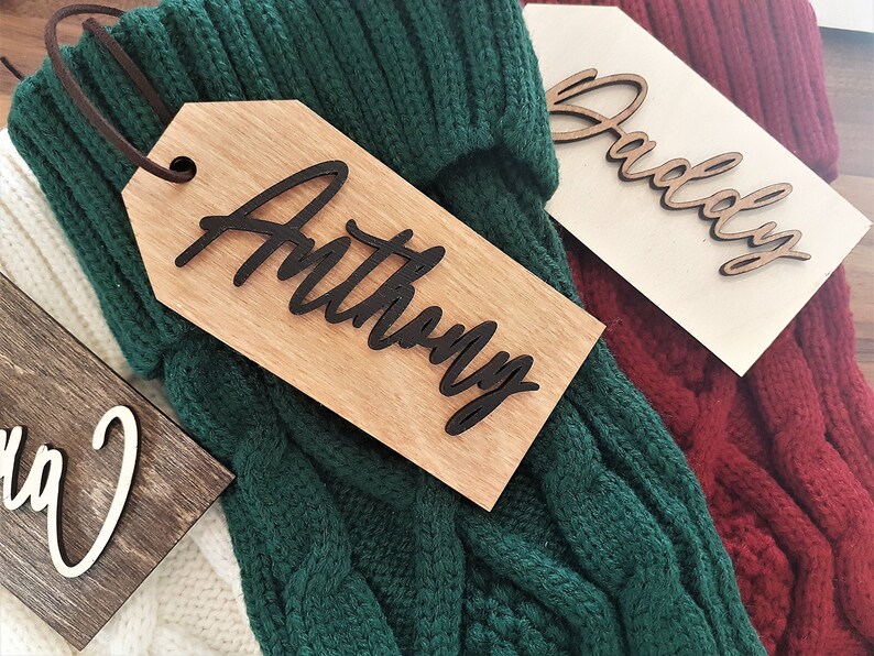 May include: Three wooden gift tags with names written in black script. The tags are attached to green, white, and red knitted stockings.