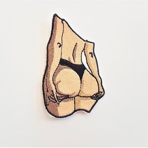 Puede incluir: Parche bordado de la espalda de una persona en la parte inferior de un bikini negro. El parche es beige y marr&oacute;n con costuras negras.