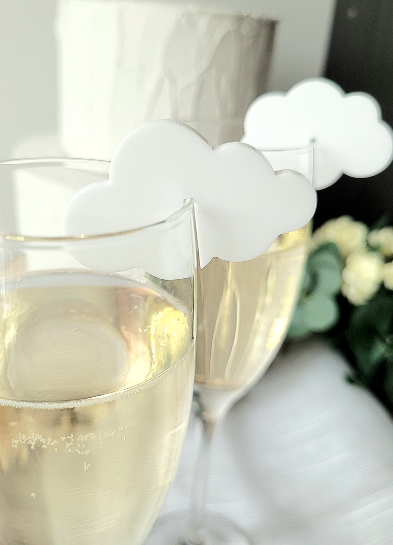 Peut inclure: Deux fl&ucirc;tes &agrave; champagne en verre transparent avec des marqueurs de boisson en forme de nuage blanc. Les fl&ucirc;tes sont remplies d'une boisson p&eacute;tillante jaune p&acirc;le.