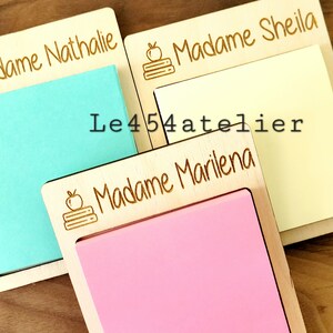 Peut inclure: Trois blocs-notes en bois avec des noms personnalis&eacute;s, "Madame Nathalie", "Madame Sheila" et "Madame Marilena". Chaque bloc-notes a un petit dessin d'une pomme et d'une pile de livres grav&eacute; sur le devant. Les blocs-notes sont sur une surface en bois.