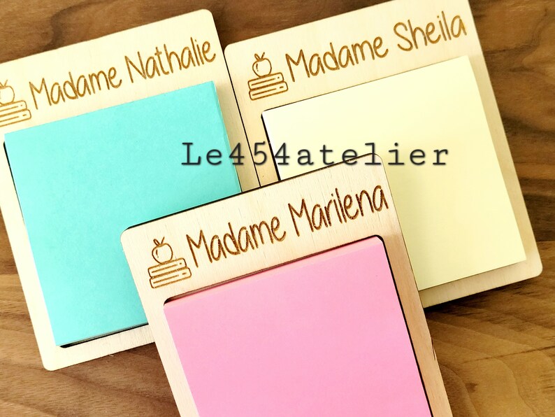 Peut inclure: Trois blocs-notes en bois avec des noms personnalis&eacute;s, "Madame Nathalie", "Madame Sheila" et "Madame Marilena". Chaque bloc-notes a un petit dessin d'une pomme et d'une pile de livres grav&eacute; sur le devant. Les blocs-notes sont sur une surface en bois.