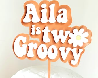 Peut inclure: Un topper de gâteau "Aila is two groovy" avec une marguerite blanche et un fond orange. Le texte est en blanc avec une police rétro.