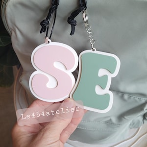 Peut inclure: Deux lettres surdimensionnées colorées, "S" et "E", sont attachées à un cordon noir et une chaîne argentée. Le "S" est rose et le "E" est vert. Les lettres sont suspendues à un sac à dos gris. Le texte "Le454atelier" est visible sur l'image.