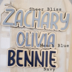 May include: Three wooden name signs with painted letters. The top sign says "Zachary" in gray paint. The middle sign says "Olivia" in gray paint. The bottom sign says "Bennie" in navy blue paint.