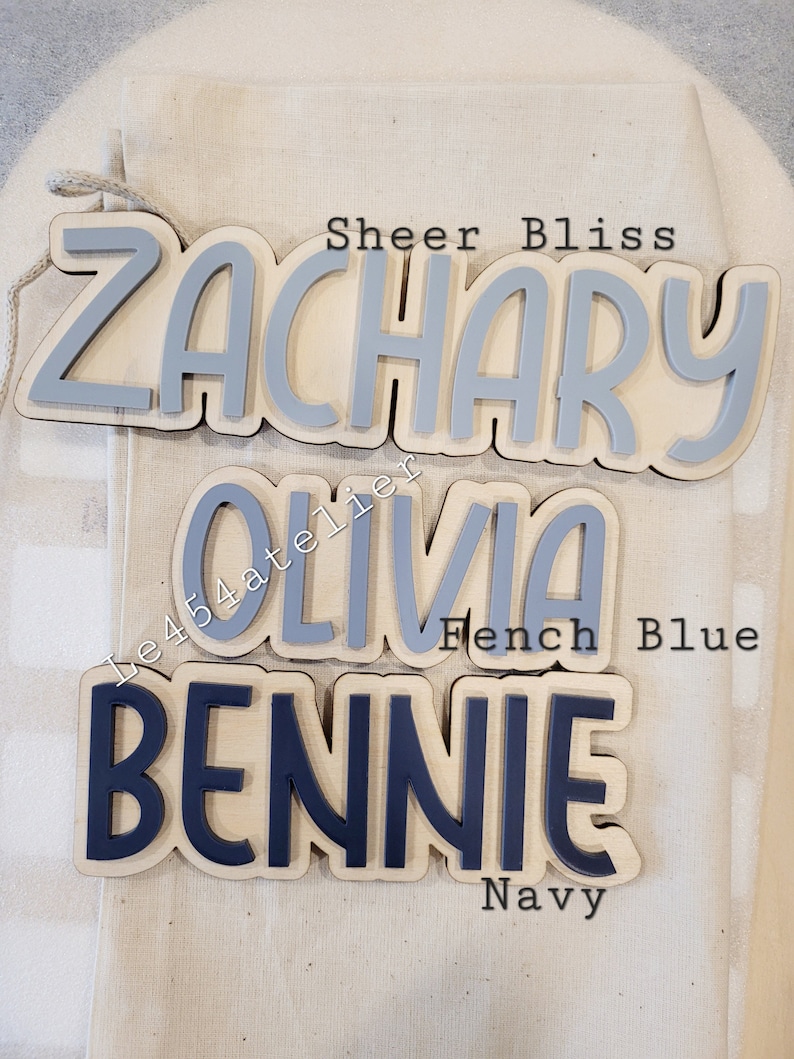 May include: Three wooden name signs with painted letters. The top sign says "Zachary" in gray paint. The middle sign says "Olivia" in gray paint. The bottom sign says "Bennie" in navy blue paint.