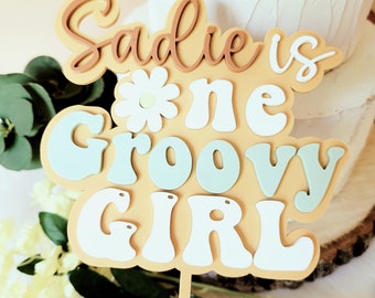 Peut inclure: Un topper de gâteau jaune, blanc et bleu avec le texte "Sadie is one groovy girl" dans une police rétro. Une marguerite blanche avec un centre jaune est au-dessus du mot "one".