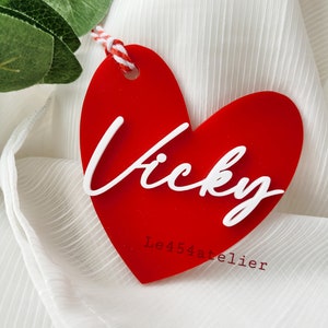 K&ouml;nnte beinhalten: Roter herzf&ouml;rmiger Acrylanh&auml;nger mit dem Namen "Vicky" in wei&szlig;er, kursiv geschriebener Schrift. Der Anh&auml;nger ist an einer wei&szlig;en und roten Schnur befestigt und tr&auml;gt den Text "Le454atelier" in wei&szlig; unter dem Namen.