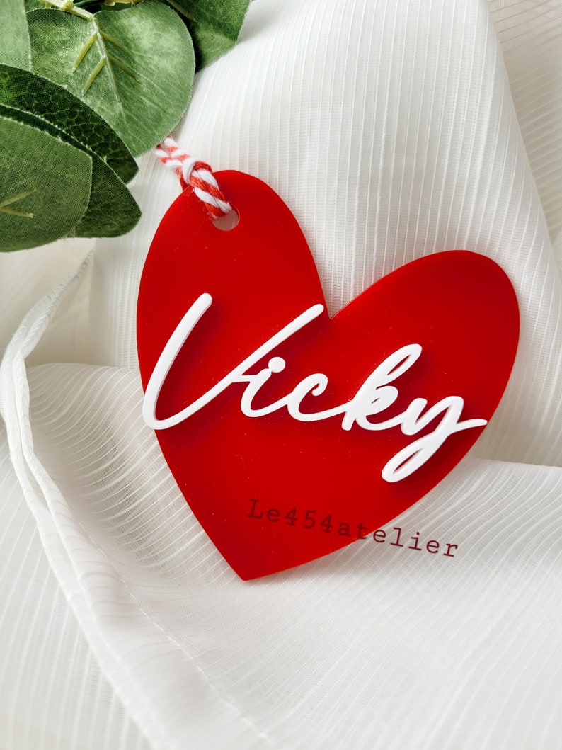 K&ouml;nnte beinhalten: Roter herzf&ouml;rmiger Acrylanh&auml;nger mit dem Namen "Vicky" in wei&szlig;er, kursiv geschriebener Schrift. Der Anh&auml;nger ist an einer wei&szlig;en und roten Schnur befestigt und tr&auml;gt den Text "Le454atelier" in wei&szlig; unter dem Namen.