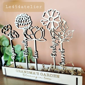 Peut inclure: Ensemble de bâtonnets de fleurs en bois avec les mots "Grandma's Garden, Blooming in Love" sur une base en bois. Les fleurs sont découpées en forme de marguerite, de lotus, de trèfle et de houx.