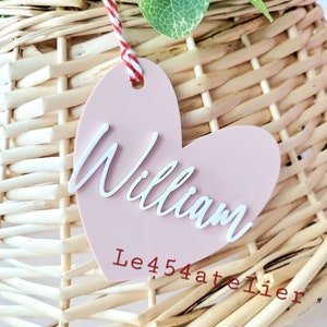 K&ouml;nnte beinhalten: Ein rosa herzf&ouml;rmiges Etikett mit dem Namen "William" in wei&szlig;er, kursiv geschriebener Schrift. Das Etikett h&auml;ngt an einem Weidenkorb mit einem roten und wei&szlig;en Bindfaden. Der Text "Le454atelier" ist in rot unter dem Namen geschrieben.