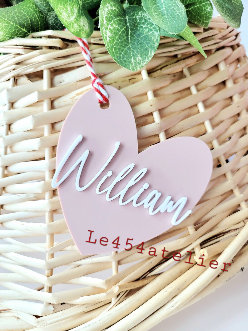 K&ouml;nnte beinhalten: Ein rosa herzf&ouml;rmiges Etikett mit dem Namen "William" in wei&szlig;er, kursiv geschriebener Schrift. Das Etikett h&auml;ngt an einem Weidenkorb mit einem roten und wei&szlig;en Bindfaden. Der Text "Le454atelier" ist in rot unter dem Namen geschrieben.