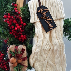 May include: White knitted Christmas stocking with a wooden gift tag that says "Dayton" in gold lettering. The stocking is hanging on a wooden ladder with a small gift box and red berries.