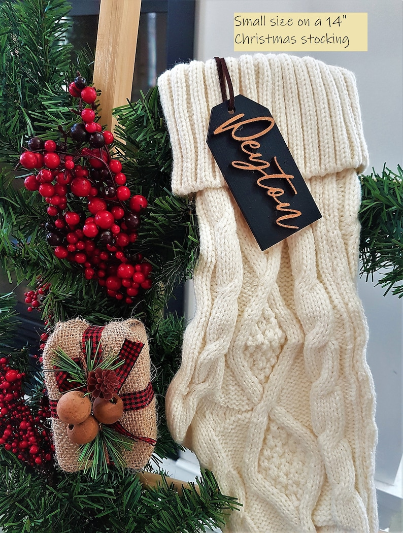 May include: White knitted Christmas stocking with a wooden gift tag that says "Dayton" in gold lettering. The stocking is hanging on a wooden ladder with a small gift box and red berries.