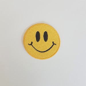 Peut inclure: Un écusson brodé jaune en forme de cercle avec un visage souriant. Le visage souriant a deux yeux ovales noirs et une bouche noire incurvée. L'écusson est sur fond blanc.