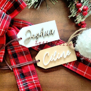 May include: Two wooden gift tags with names written in white cursive lettering. The tag on the left says "Joshua" and the tag on the right says "Celine". The tags are tied together with a brown cord and are sitting on a red and black plaid ribbon.