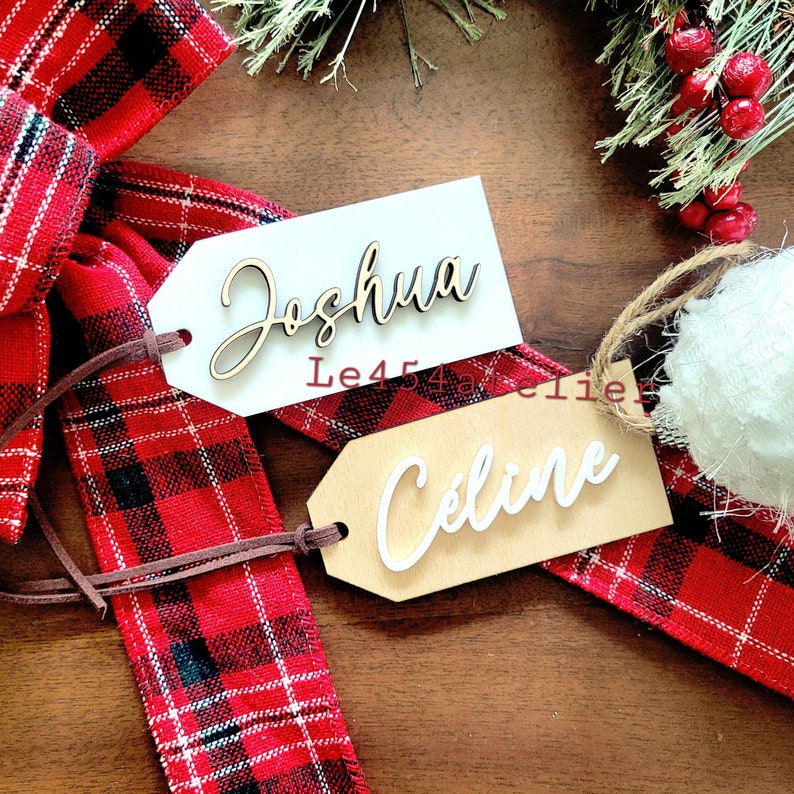 May include: Two wooden gift tags with names written in white cursive lettering. The tag on the left says "Joshua" and the tag on the right says "Celine". The tags are tied together with a brown cord and are sitting on a red and black plaid ribbon.