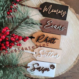 Peut inclure: Cinq étiquettes cadeaux en bois avec des noms écrits en cursive. Les étiquettes sont dans différentes nuances de brun et de blanc. Les noms sur les étiquettes sont "Mommy", "Evans", "Rhett", "Sutton" et "Snipe".