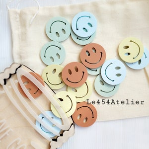 以下が含まれることがあります： 白い布製のバッグに、カラフルなスマイリーフェイスのボタンがたくさん入っています。ボタンは青、緑、黄、オレンジの色合いです。バッグには「Le454Atelier」という文字が見えます。