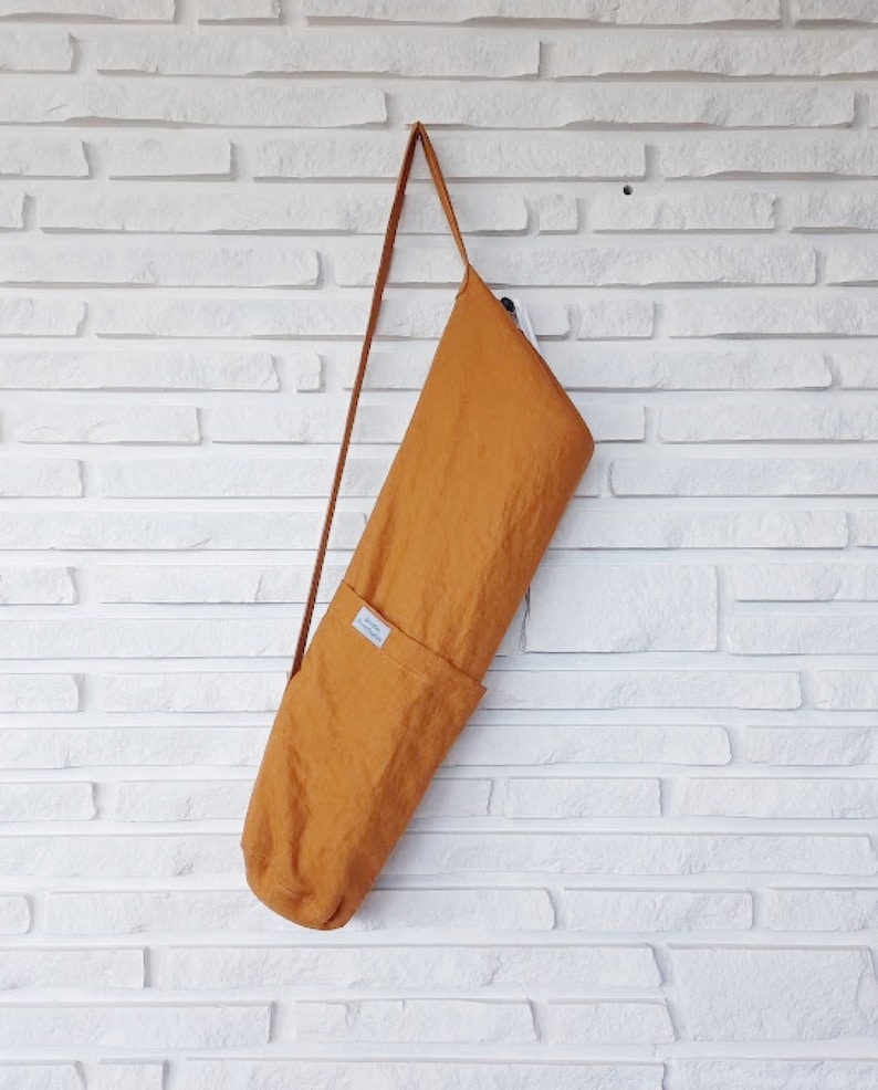 May include: Mustard-colored yoga mat bag with a long, thin strap. The bag is made of a textured fabric and has a small pocket on the front. It hangs against a white brick wall.