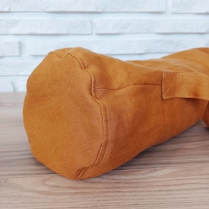 May include: A cylindrical, burnt orange yoga mat bag made of a textured fabric. The bag has a rectangular handle on the side and is designed to carry a rolled-up yoga mat. The bag is sitting on a wooden surface.