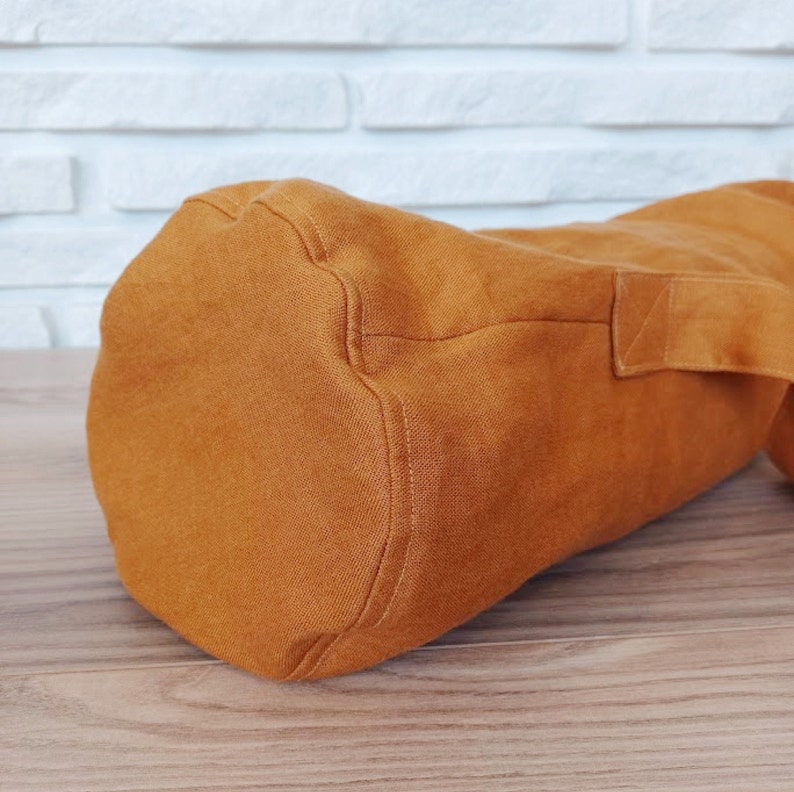 May include: A cylindrical, burnt orange yoga mat bag made of a textured fabric. The bag has a rectangular handle on the side and is designed to carry a rolled-up yoga mat. The bag is sitting on a wooden surface.