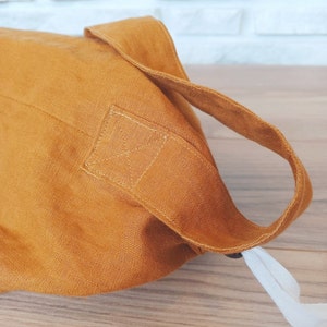 May include: Mustard-colored linen tote bag with a sturdy handle and a white drawstring closure. The bag's handle is attached with reinforced stitching. The bag is made of a textured fabric.