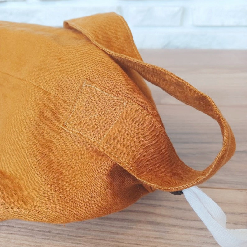May include: Mustard-colored linen tote bag with a sturdy handle and a white drawstring closure. The bag's handle is attached with reinforced stitching. The bag is made of a textured fabric.