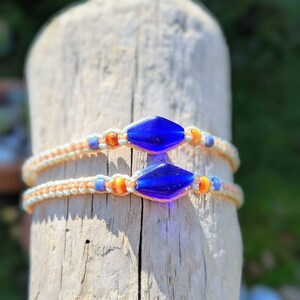 Puede incluir: Dos pulseras con cuerda blanca y naranja, y cuentas de vidrio azul. Las pulseras están atadas alrededor de un trozo de madera flotante.