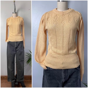 May include: A pale yellow knitted sweater with a diamond pattern and a ribbed hem. The sweater has long sleeves and a round neckline.