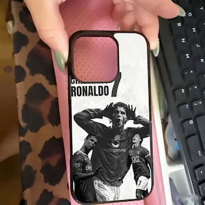 Puede incluir: Funda de tel&eacute;fono negra con im&aacute;genes en blanco y negro de Cristiano Ronaldo con la camiseta del Manchester United. El nombre "RONALDO" est&aacute; impreso en la parte superior. Tambi&eacute;n es visible el n&uacute;mero "7".